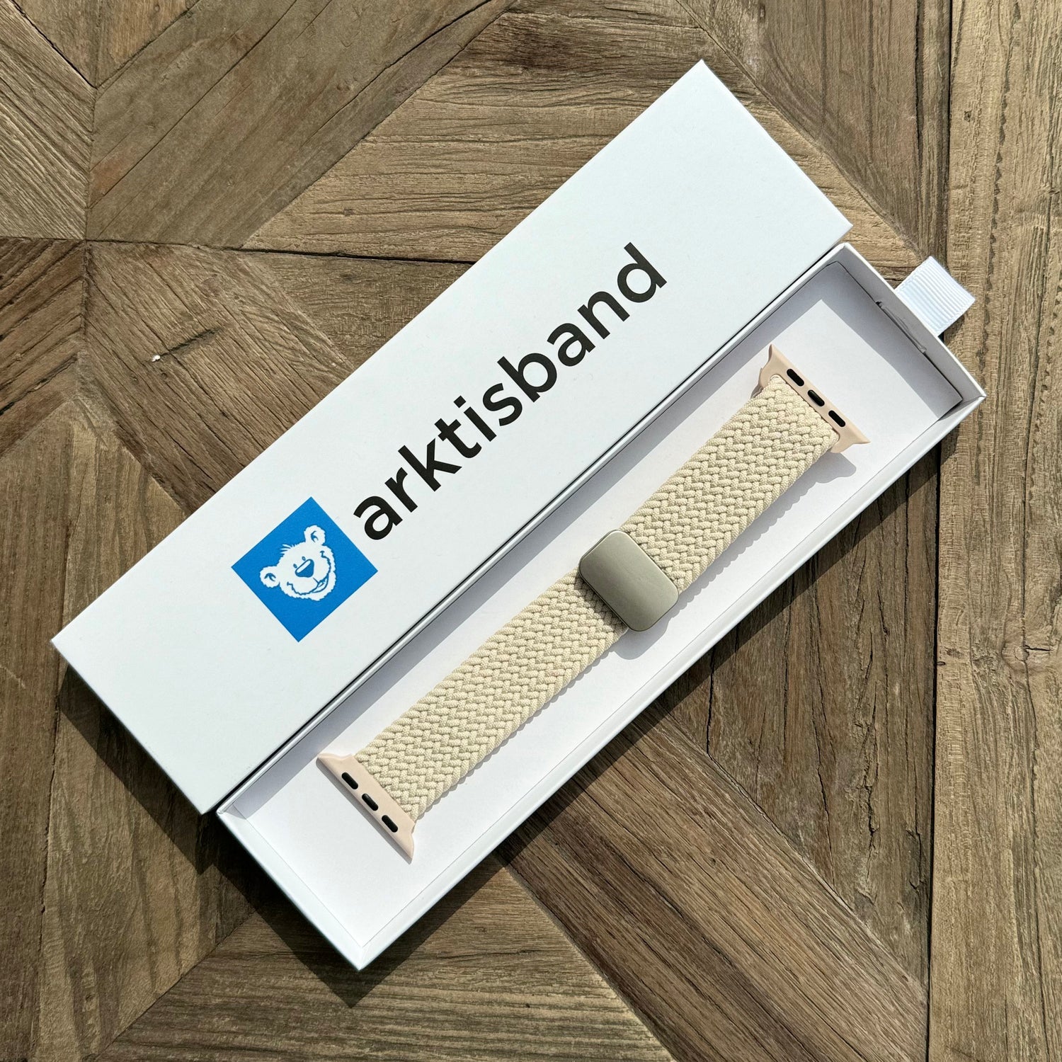 Ein von Arktisband geflochtenes Apple Watch-Armband „Casual“ mit Magnetverschluss in Creme wird in einer offenen weißen Arktisband-Box mit einem blauen Eisbär-Logo auf einer hölzernen, geometrisch gemusterten Oberfläche gezeigt.