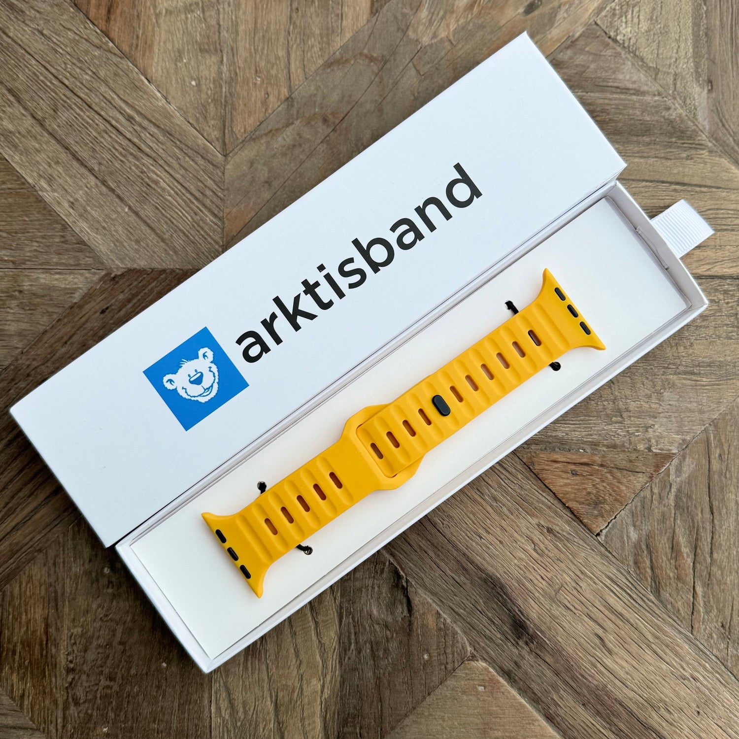 Ein gelbes Arktisband Apple Watch Silikonarmband „Active“ mit atmungsaktivem Design wird in einer offenen weißen Arktisband-Box mit einem blauen Bärengesichtslogo auf einer Holzoberfläche gezeigt.