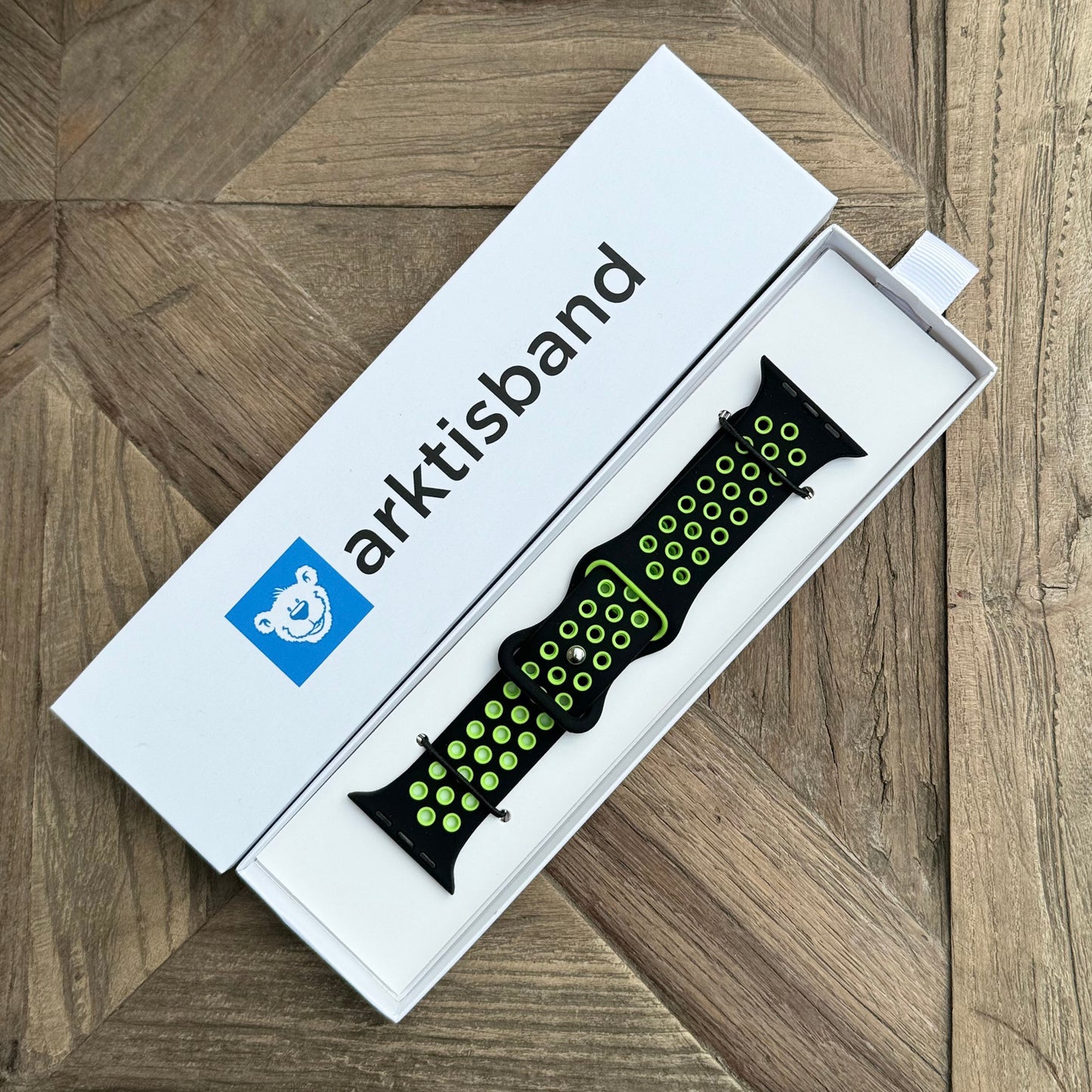 Das arktisband Apple Watch Silikonarmband „Energy“, ein schwarzes Band mit grünen Löchern, wird in seiner weißen Arktisband-Box mit blauem Bären-Logo präsentiert. Die Box ist ideal als Sporttrainingszubehör und liegt auf einer Holzoberfläche mit geometrischem Muster.