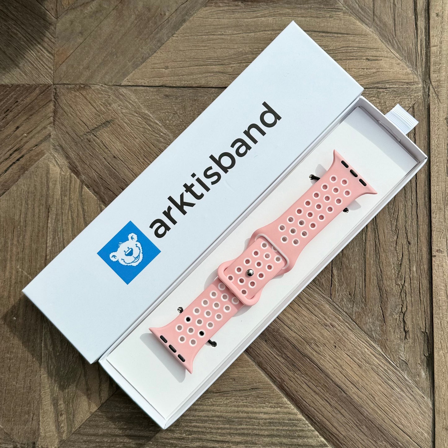 Ein Arktisband Apple Watch Silikonarmband „Energy“ in Pink und Weiß wird in einer offenen weißen Arktisband-Box auf einer Holzoberfläche gezeigt.