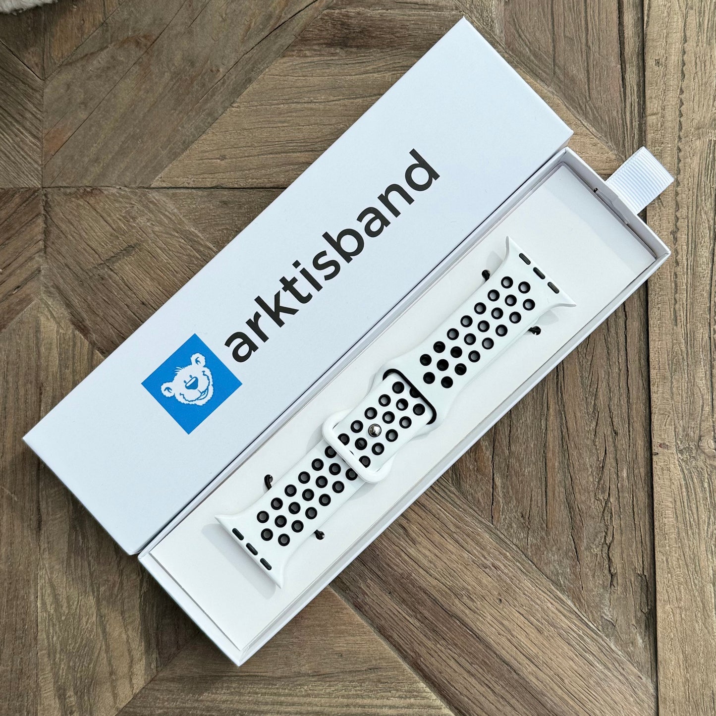 Das Arktisband Apple Watch Silikonarmband „Energy“ in Weiß mit schwarzen Löchern wird in einer weißen rechteckigen Arktisband-Box mit blauem Bärenlogo auf einer geometrisch gemusterten Holzfläche präsentiert.