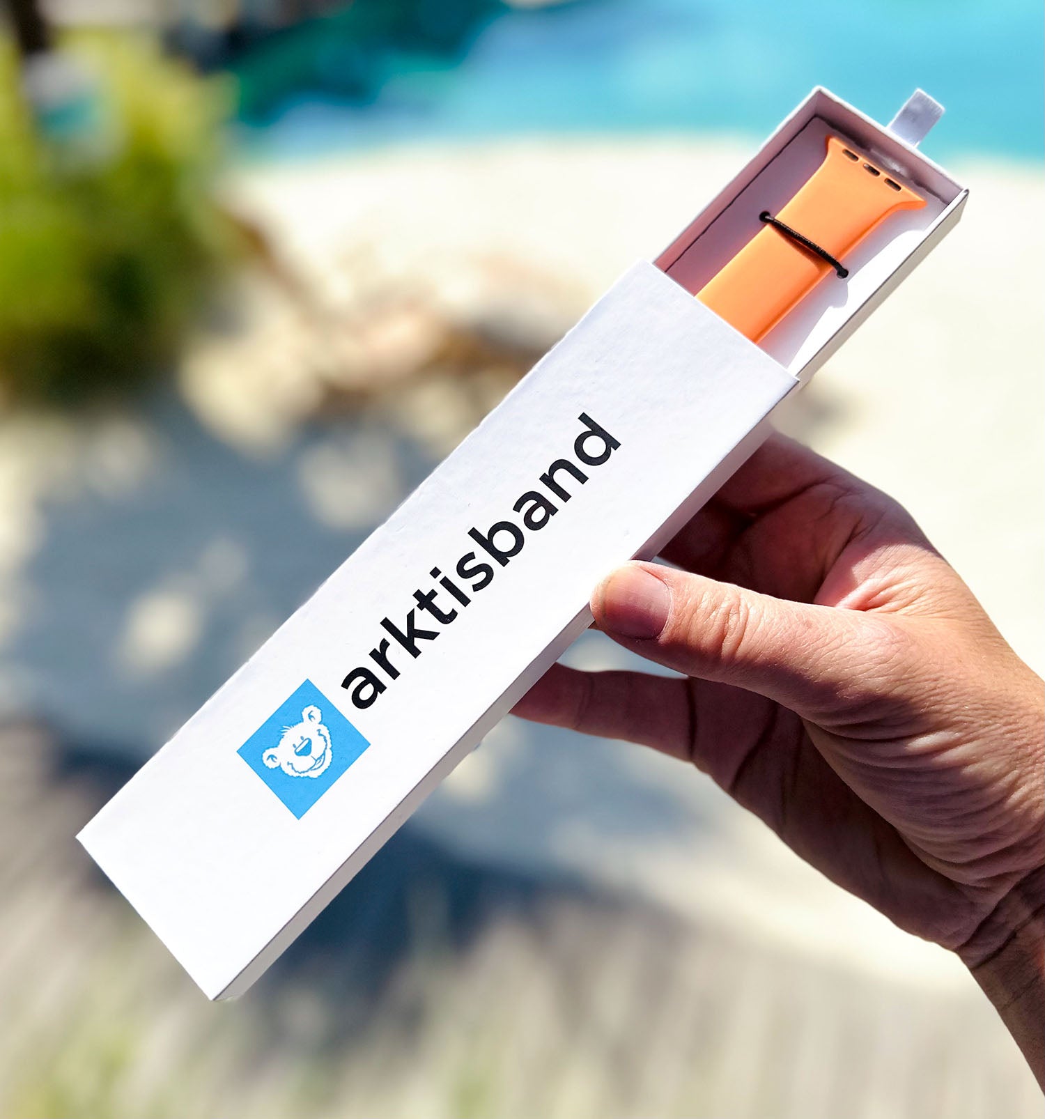 Eine Hand hält eine Arktisband-Box mit einem orangefarbenen Smartwatch-Armband in der Nähe eines Pools.