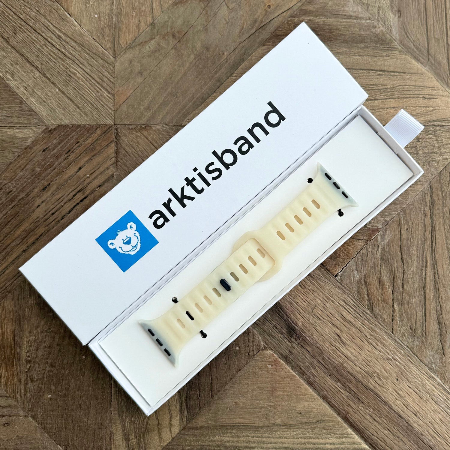 Ein Arktisband Apple Watch Silikonarmband „Active“ in Weiß mit schwarzen Akzenten wird in seiner offenen Box mit dem blauen Bärenlogo auf einer Oberfläche mit Holzmuster gezeigt.
