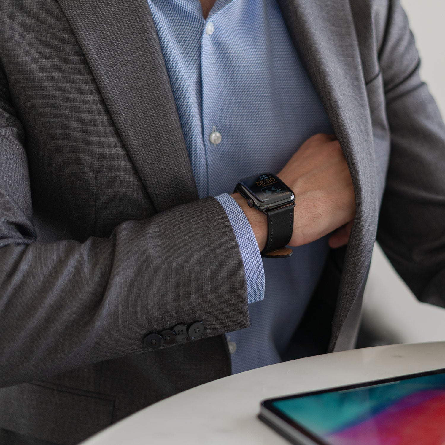 Mann im Anzug mit Smartwatch, Hand in der Jacke, Tablet auf einem Tisch davor.