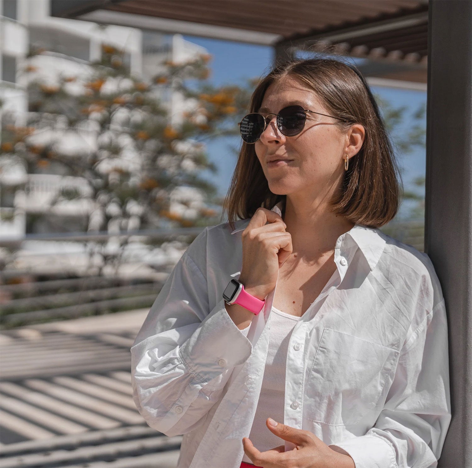 Frau mit Sonnenbrille und weißem Hemd steht im Freien, trägt eine Smartwatch mit rosa Armband und lächelt leicht.