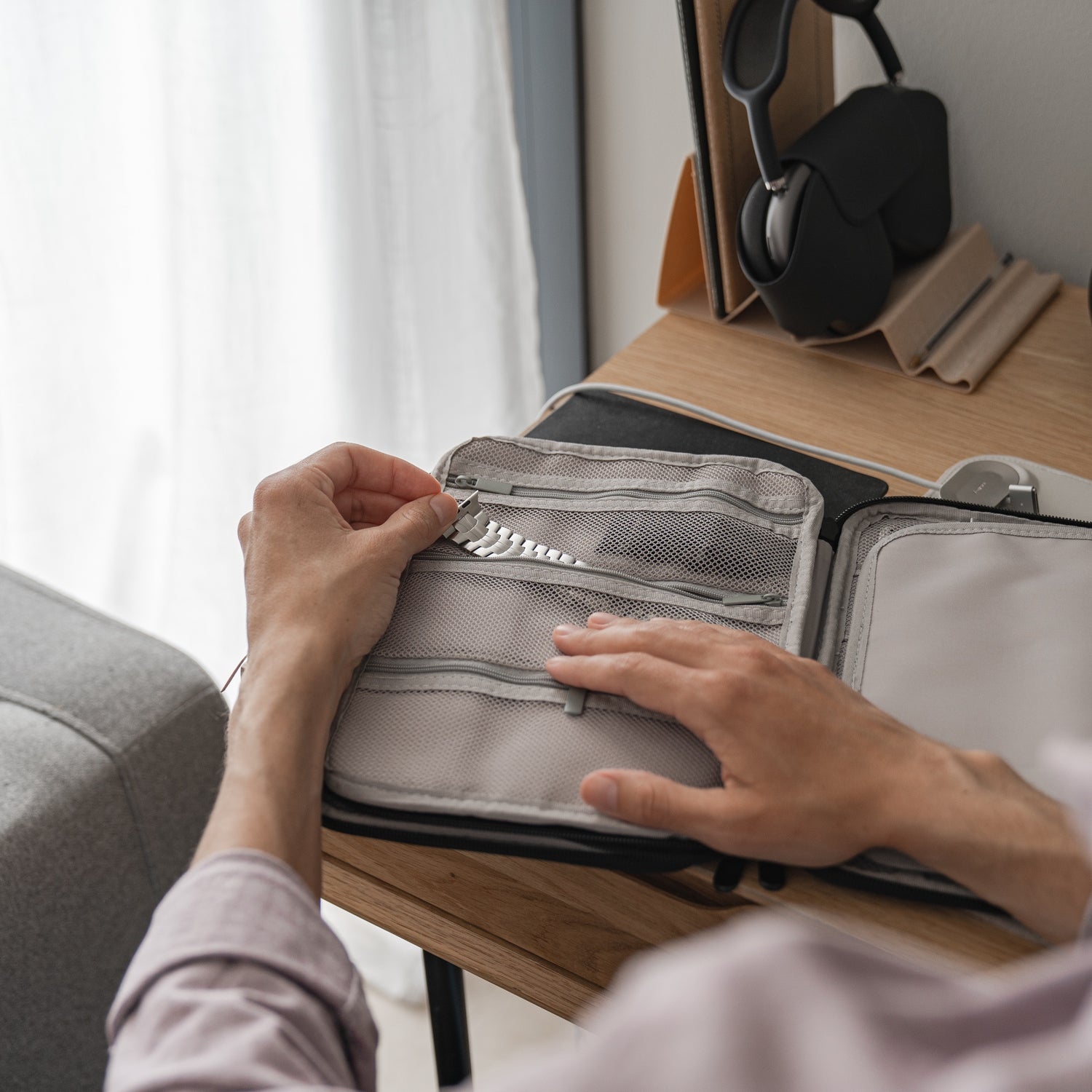 Jemand steckt eine Blisterpackung mit Pillen in die Netztasche einer kleinen Reisetasche für Apple Watch Armbänder & Zubehör von arktisband auf einem Holzschreibtisch neben Kopfhörern, anderem Zubehör und einer Couch.