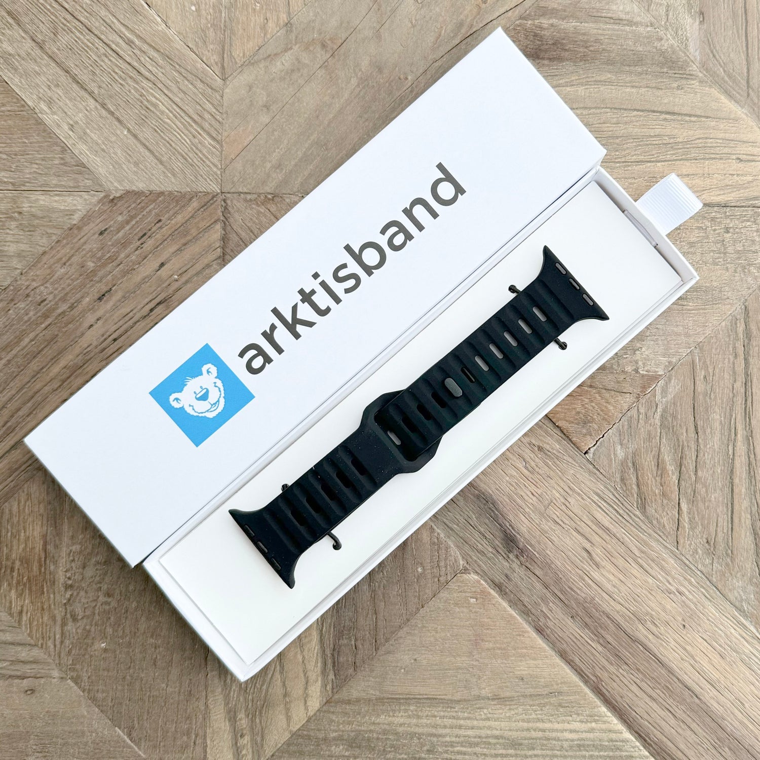 Das arktisband Apple Watch Silikonarmband "Active" in Schwarz präsentiert sich in einer offenen weißen arktisband-Box mit blauem Eisbär-Logo auf einer Oberfläche mit Holzmuster. Sein atmungsaktives Design bietet ganztägigen Tragekomfort.
