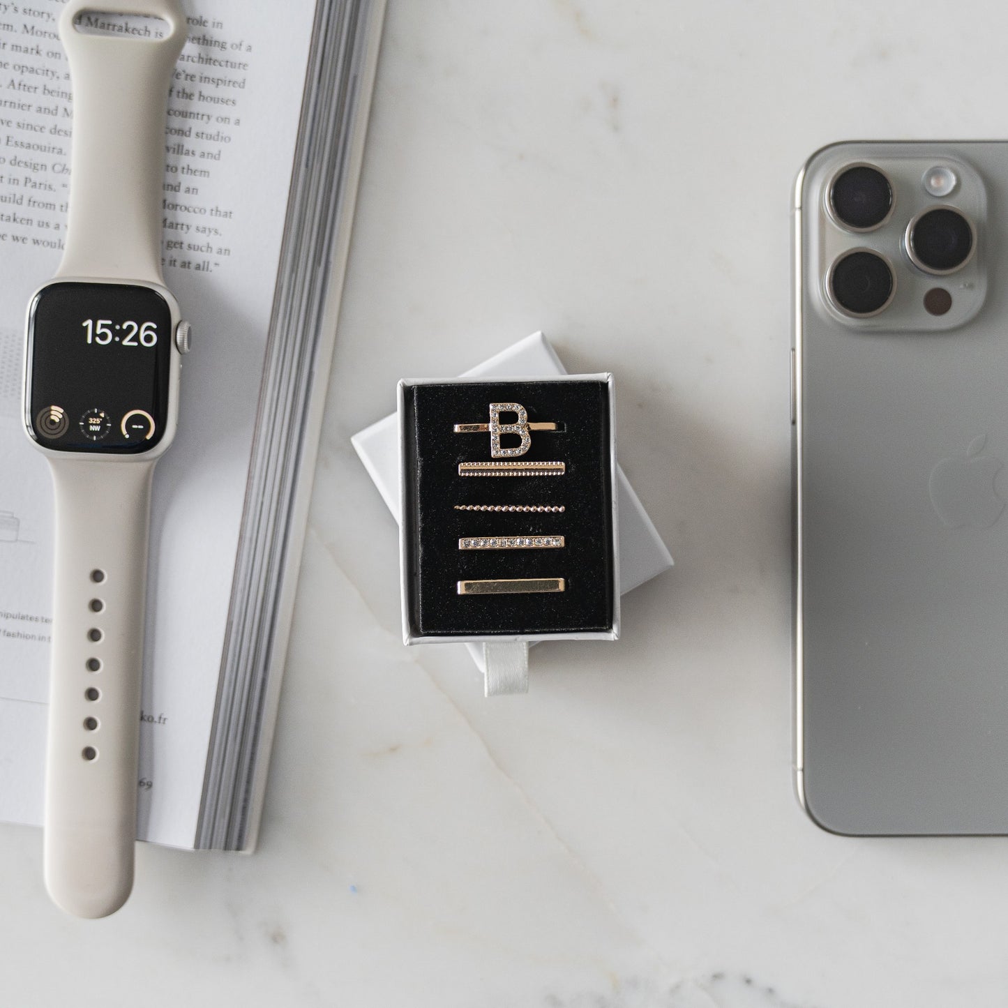 Eine beige Smartwatch mit Arktisband Apple Watch Charms „B Style“, goldene Manschettenknöpfe in einer schwarzen Box und ein silbernes Smartphone liegen auf einer weißen Marmoroberfläche neben einem offenen Buch.