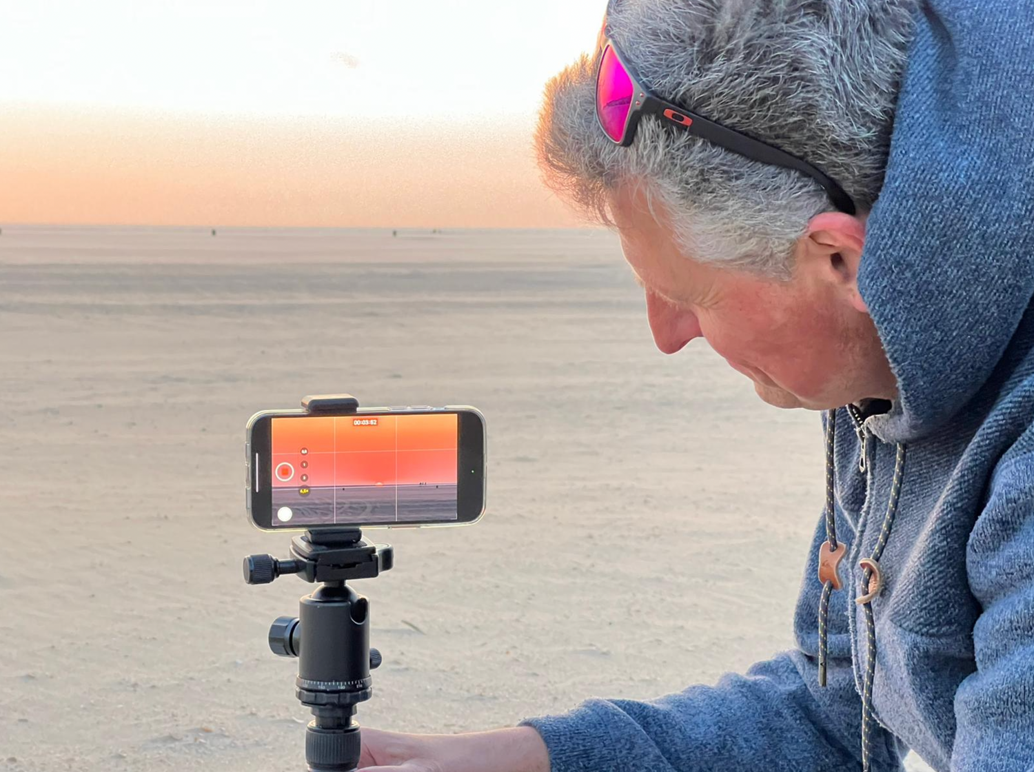 Eine Person richtet ein Smartphone auf einem Stativ aus, um einen Sonnenuntergang über einer weiten Sandlandschaft zu fotografieren.