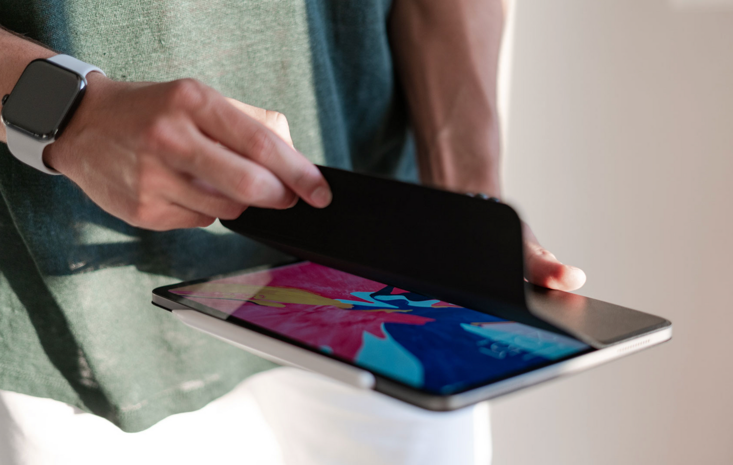Eine Person öffnet das arktis iPad Pro 11" (2024/2025) magnetisches Smart Case, zeigt einen farbenfrohen Bildschirm, trägt eine Smartwatch und ein grünes Shirt.