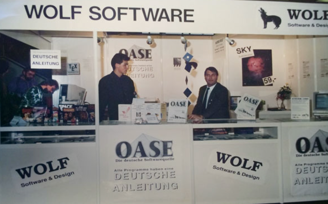 Zwei Männer stehen an einem Messestand von Wolf Software. Auf der Theke sind Schilder und Softwareboxen ausgestellt.