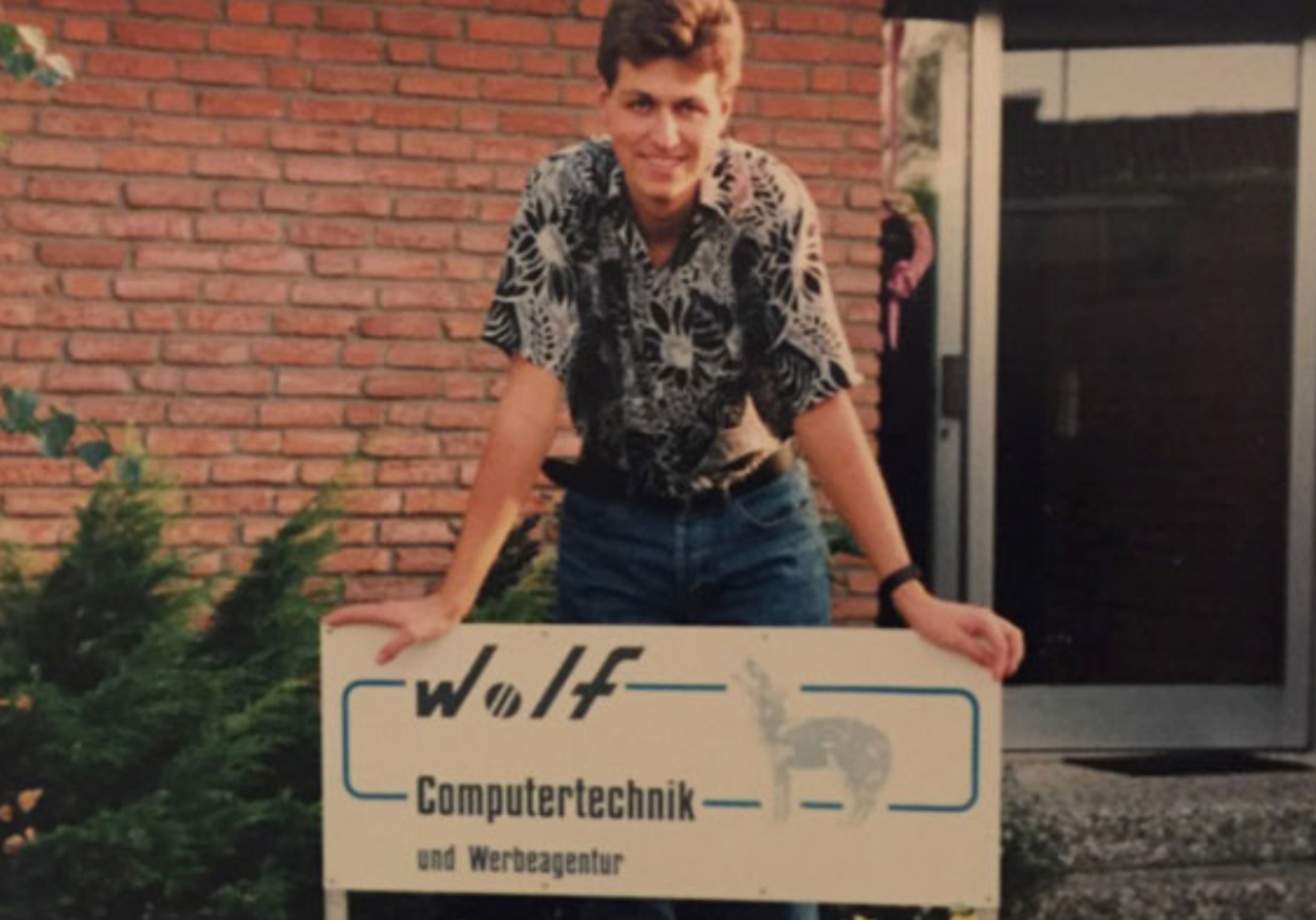 Ein Mann im gemusterten Hemd lehnt an einem Schild von Wolf Computertechnik und Werbeagentur vor einem Backsteingebäude.