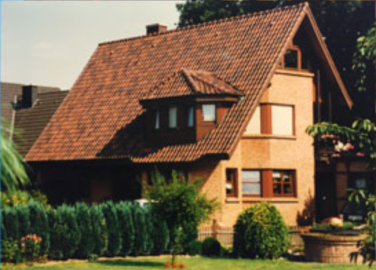 Ein zweistöckiges Backsteinhaus mit steilem Ziegeldach, braunen Fensterläden und einem Garten davor.