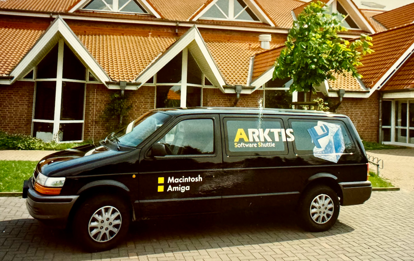 Ein schwarzer Lieferwagen mit der Aufschrift „ARKTIS Software Shuttle“ parkt vor einem Gebäude mit roten Ziegeldächern.