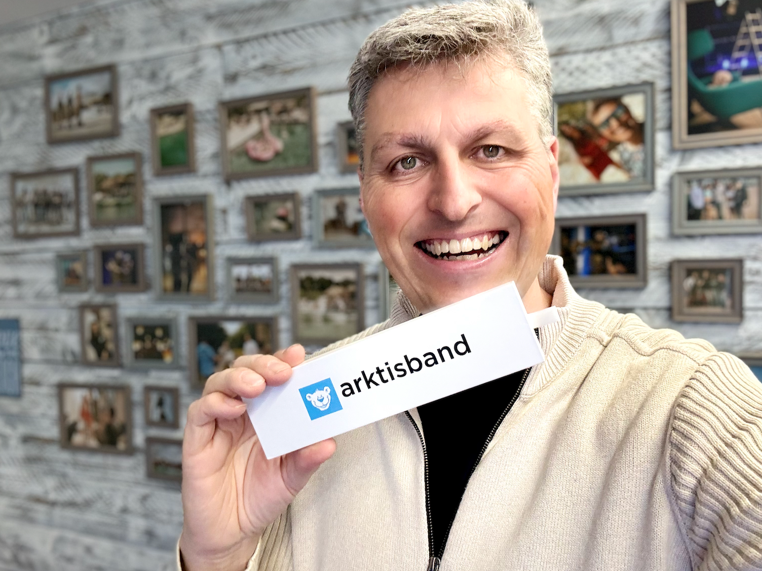 Lächelnder Mann hält ein weißes Arktisband-Schild vor einer Wand mit vielen gerahmten Fotos.