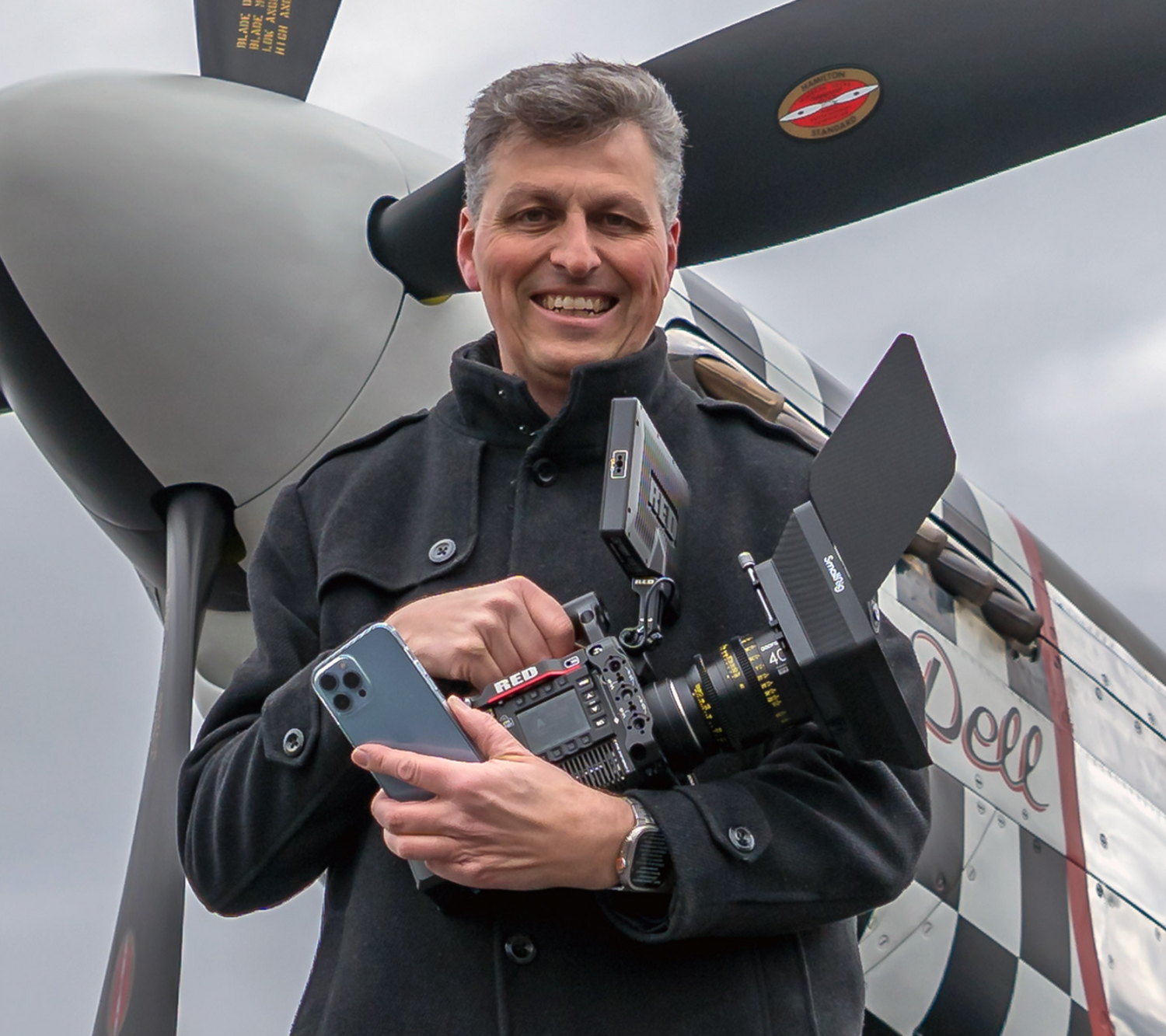 Lächelnder Mann hält eine Kamera und ein Smartphone vor einem alten Flugzeugpropeller.