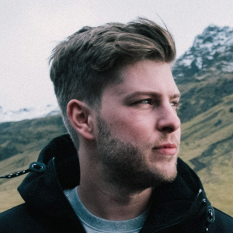 Ein Mann mit kurzen Haaren und Bart schaut zur Seite und steht im Freien in der Nähe schneebedeckter Berge.