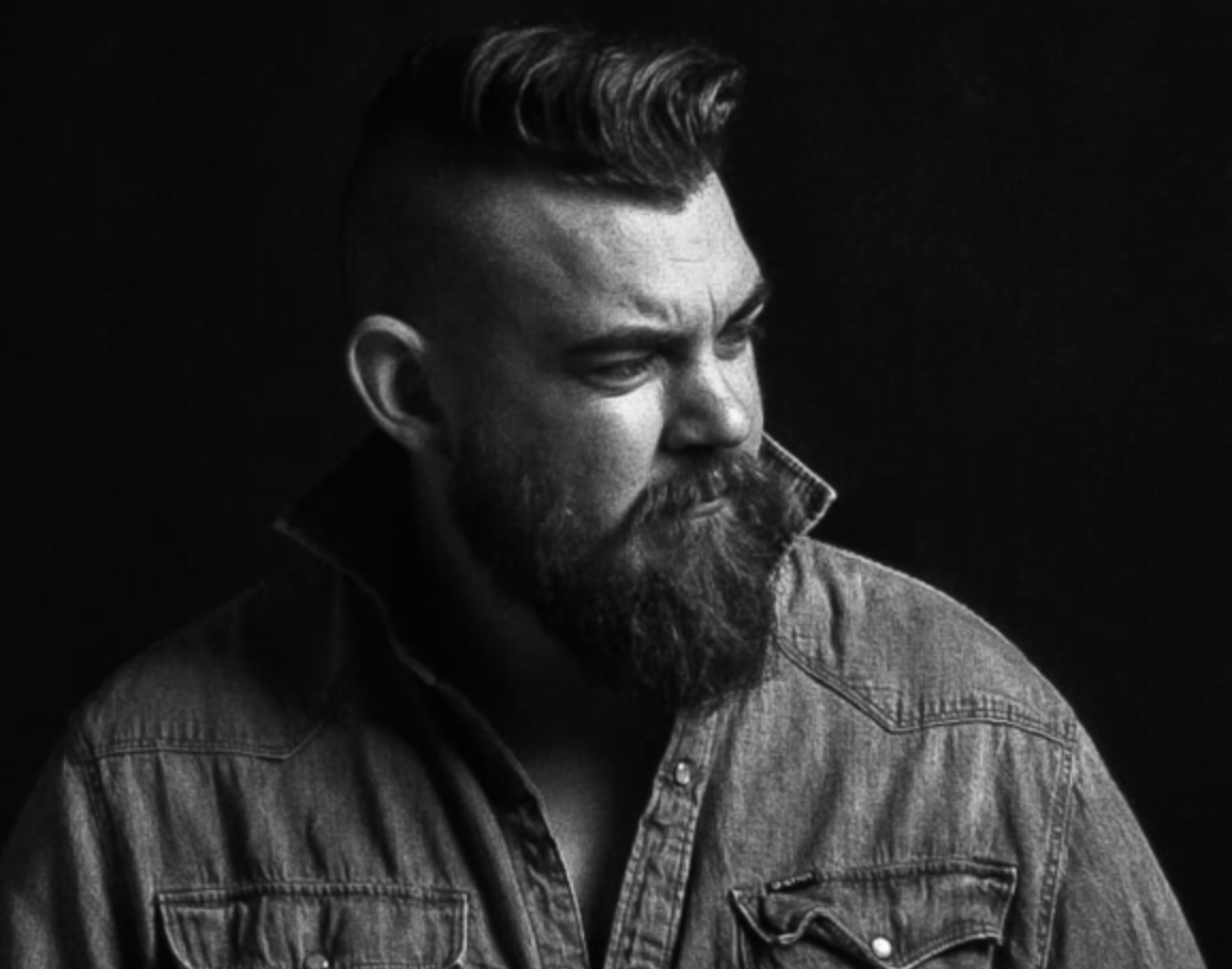 Mann mit Bart und frisiertem Haar, trägt ein Jeanshemd mit hochgestelltem Kragen und blickt zur Seite.