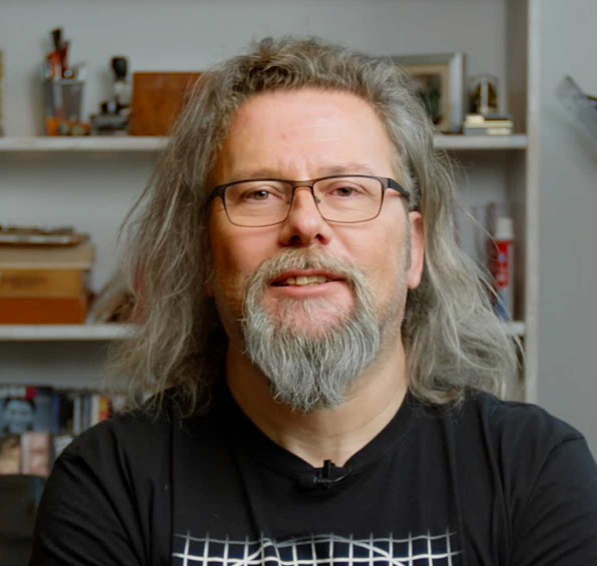 Mann mit langen grauen Haaren, Brille und Bart sitzt im Haus, im Hintergrund sind Regale und Bücher.