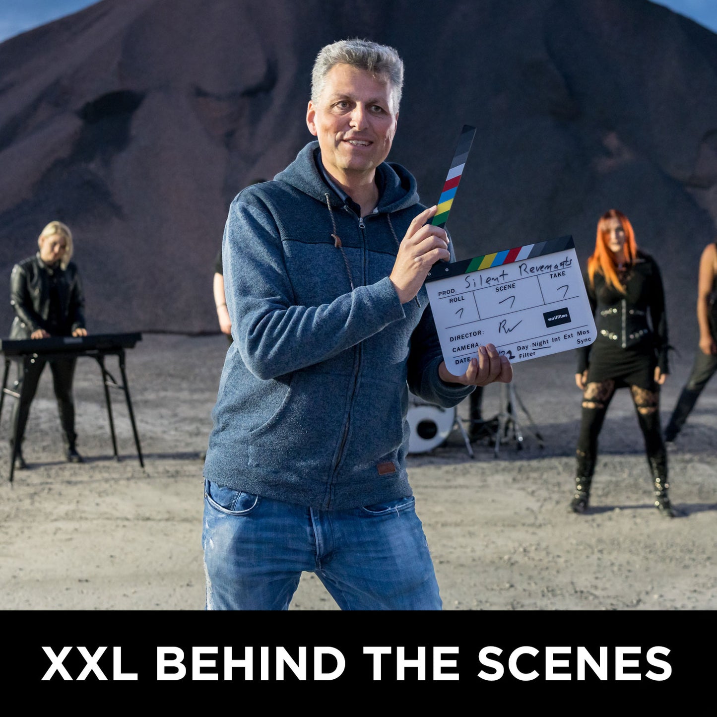 Ein Mann steht mit einer Filmklappe im Freien, hinter ihm stehen vier weitere mit Instrumenten, dazu ein Kieshaufen. Der Text lautet: XXL HINTER DEN KULISSEN: Wolffilms Academy Online-Kurs – Einfach filmen lernen von Rainer Wolf.