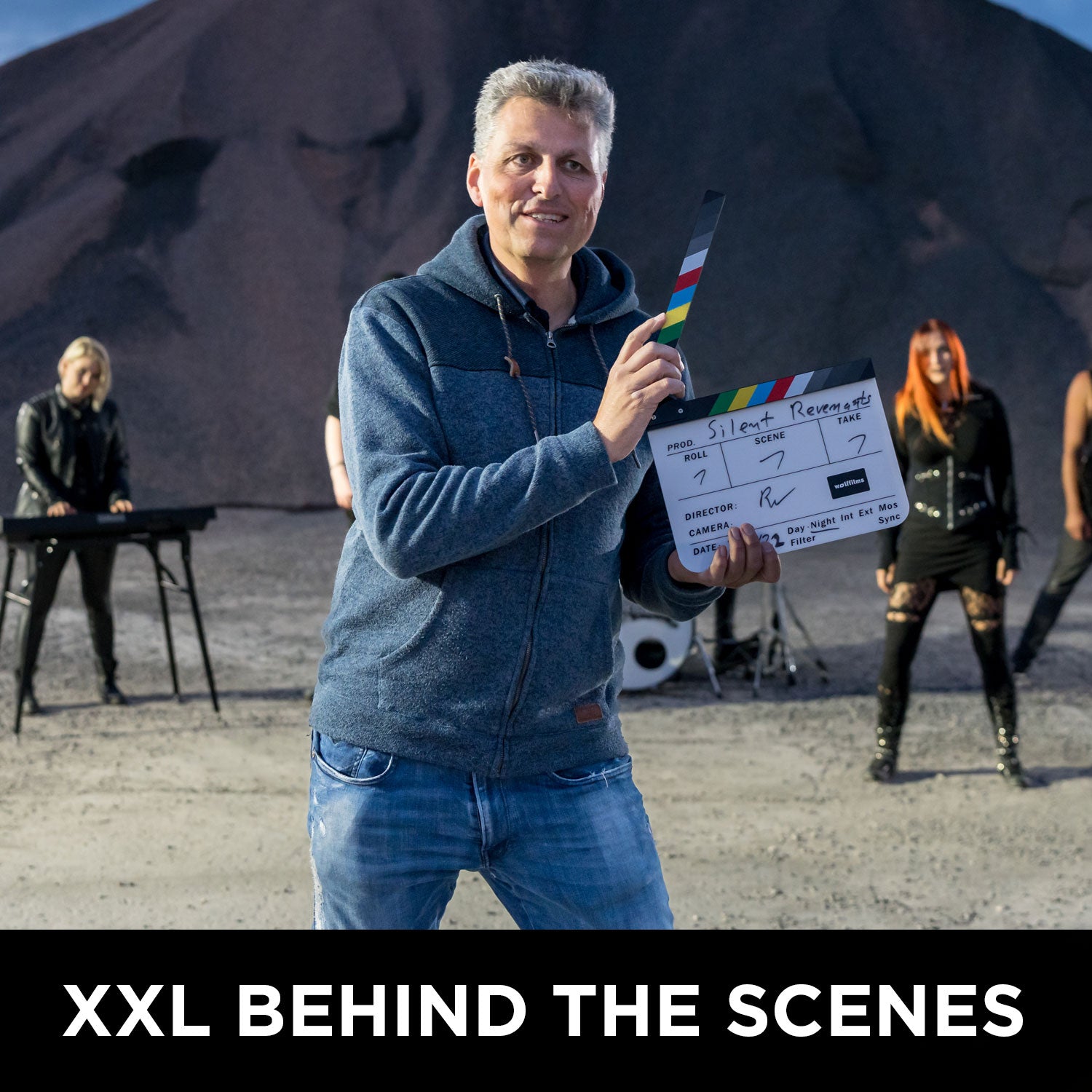Ein Mann steht mit einer Filmklappe im Freien, hinter ihm stehen vier weitere mit Instrumenten, dazu ein Kieshaufen. Der Text lautet: XXL HINTER DEN KULISSEN: Wolffilms Academy Online-Kurs – Einfach filmen lernen von Rainer Wolf.