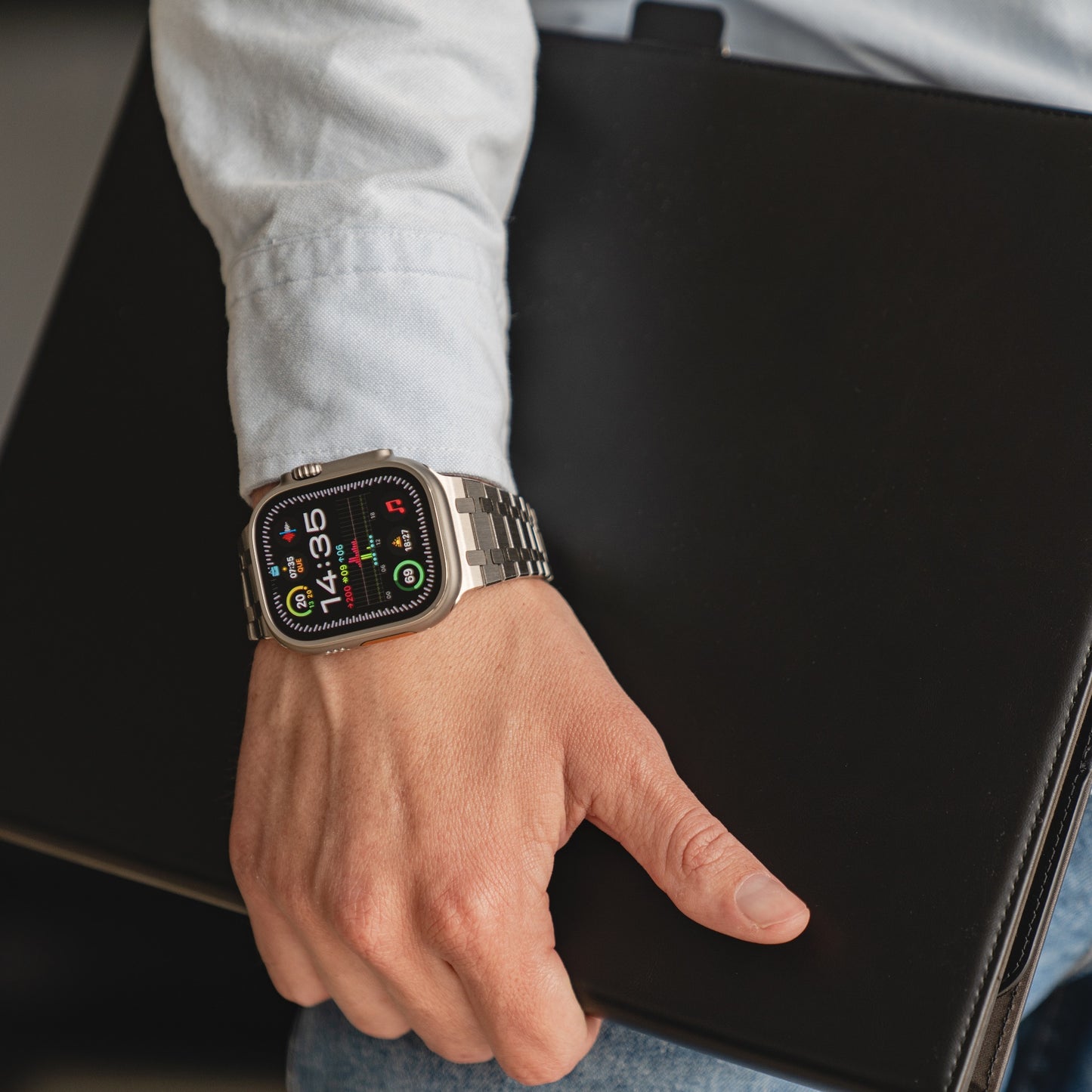Eine Person, die ein Arktisband Edelstahl Armband „Symphony“ für die Apple Watch trägt, hält einen geschlossenen schwarzen Laptop unter dem Arm.