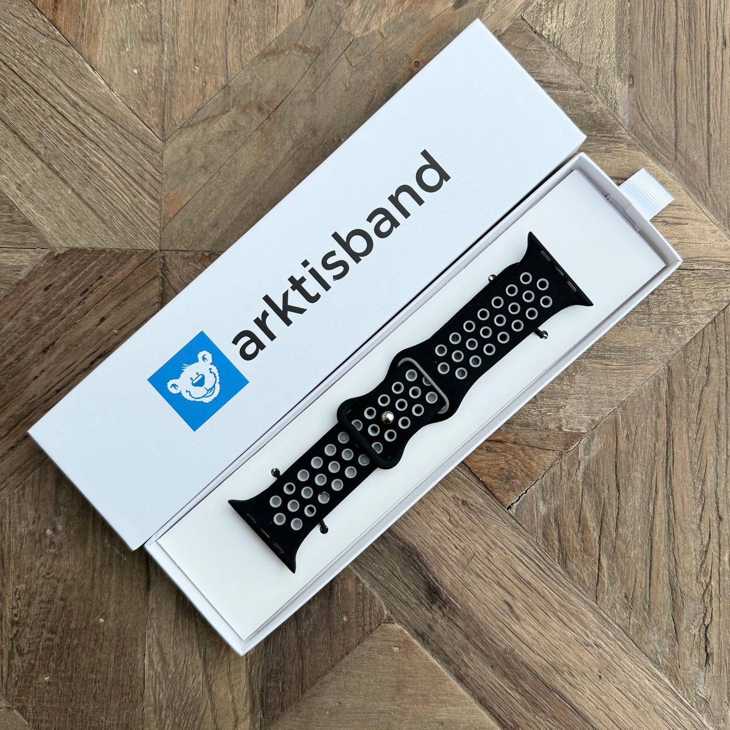 Das arktisband Apple Watch Silikonarmband „Energy“, ein schwarzes perforiertes Armband, wird in einer offenen weißen arktisband-Box mit einem blauen Eisbär-Logo auf einer Holzoberfläche mit geometrischem Muster gezeigt.