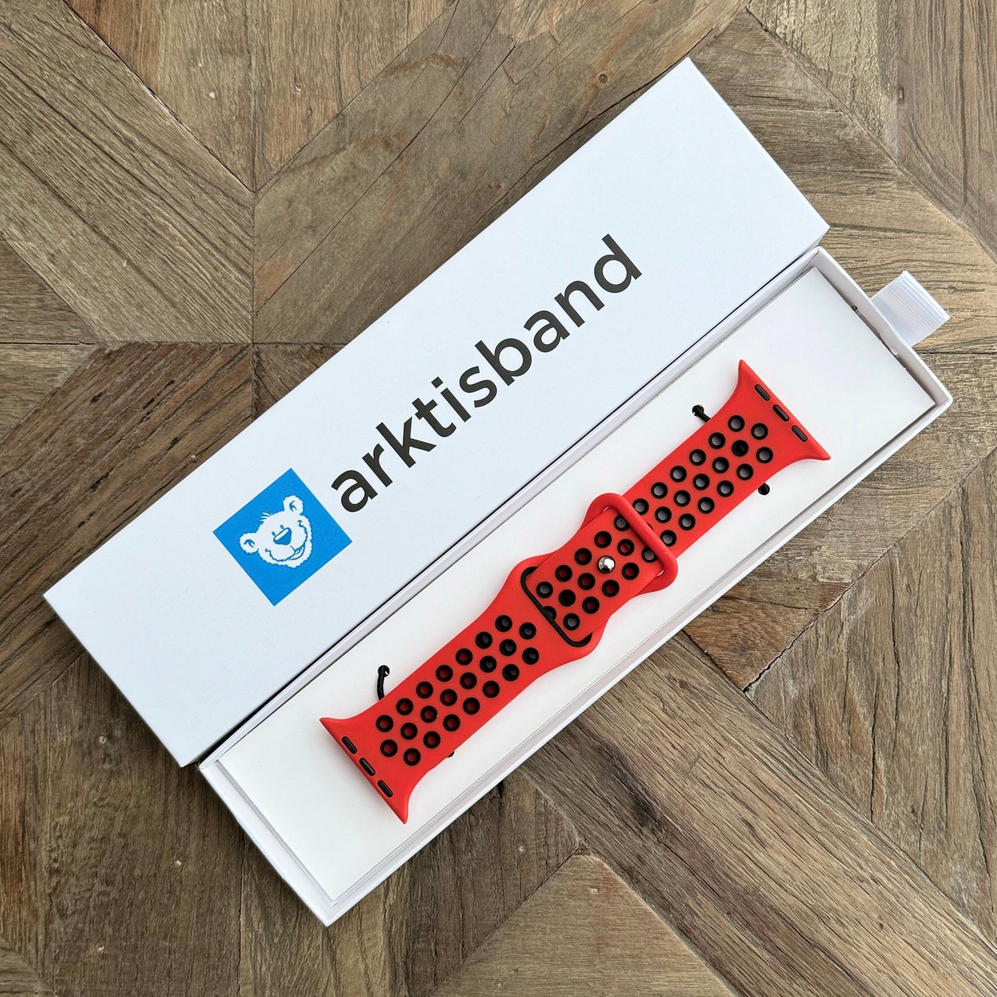 Ein Arktisband Apple Watch Silikonarmband „Energy“ in Rot und Schwarz wird in einer offenen weißen Box mit einem blauen Bärenlogo auf einer hölzernen Oberfläche mit Fischgrätenmuster gezeigt.