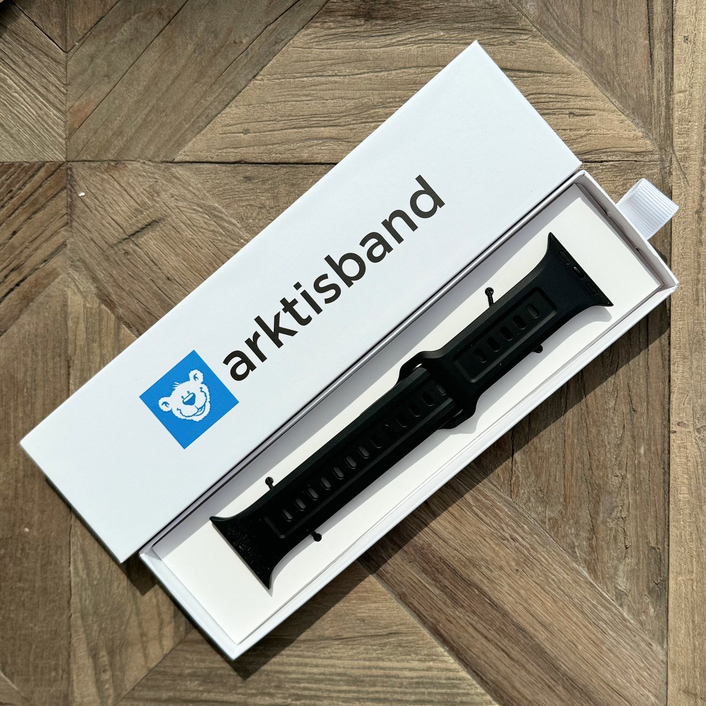 Das arktisband Apple Watch FKM Sportarmband „Sweat“ in Schwarz wird strapazierfähig und elastisch in einer weißen arktisband-Box mit blauem Eisbär-Logo auf geometrischem Holz präsentiert. Kompatibel mit Apple Watch.