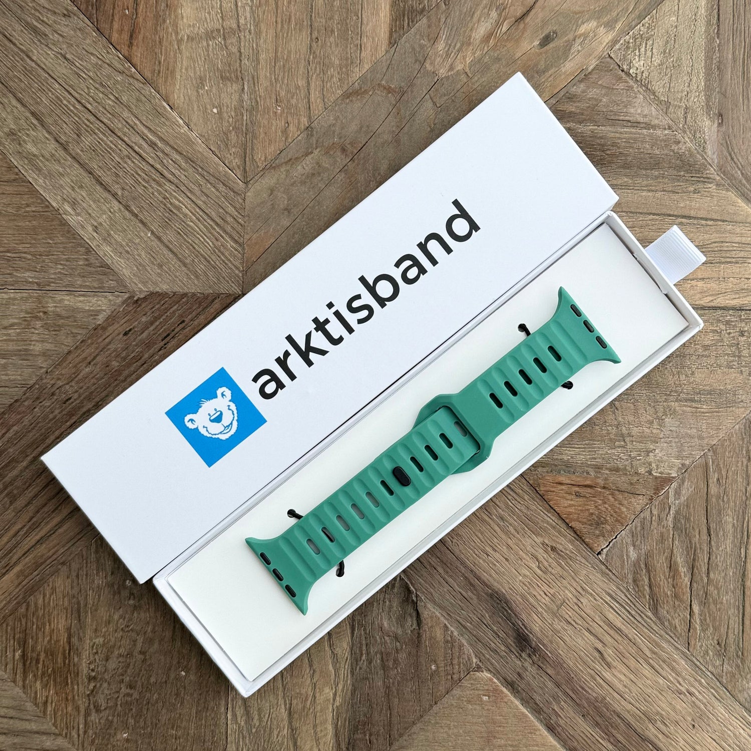 Ein arktisband Apple Watch Silikonarmband „Active“ in Grün wird in einer offenen weißen Box gezeigt, wobei das arktisband-Logo – ein blaues Quadrat mit einem weißen Bären – seinen sportlichen Look auf einer Holzoberfläche hervorhebt.