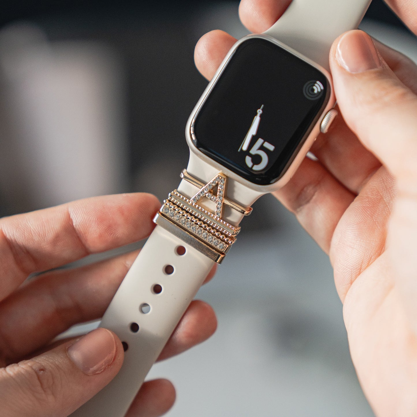 Eine Person hält eine Smartwatch mit einem beigen Armband und einem daran befestigten Arktisband Apple Watch Charms „A Style“-Anhänger, der das stylische Arktisband-Accessoire für einen personalisierten Apple Watch-Look präsentiert.