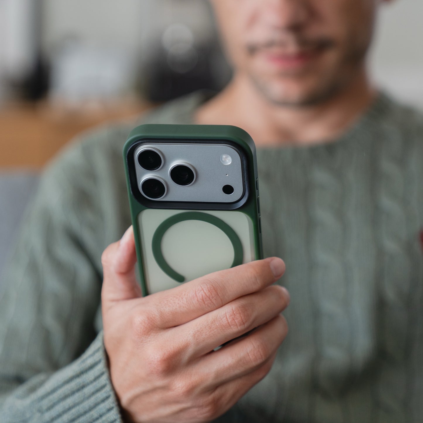 Eine Person in einem grünen Pullover hält eine Arktis iPhone 17 Pro Solid Case mit MagSafe - eine grün-weiße matte Schutzhülle von Arktis - auf ihr Smartphone und schaut in die Kamera.