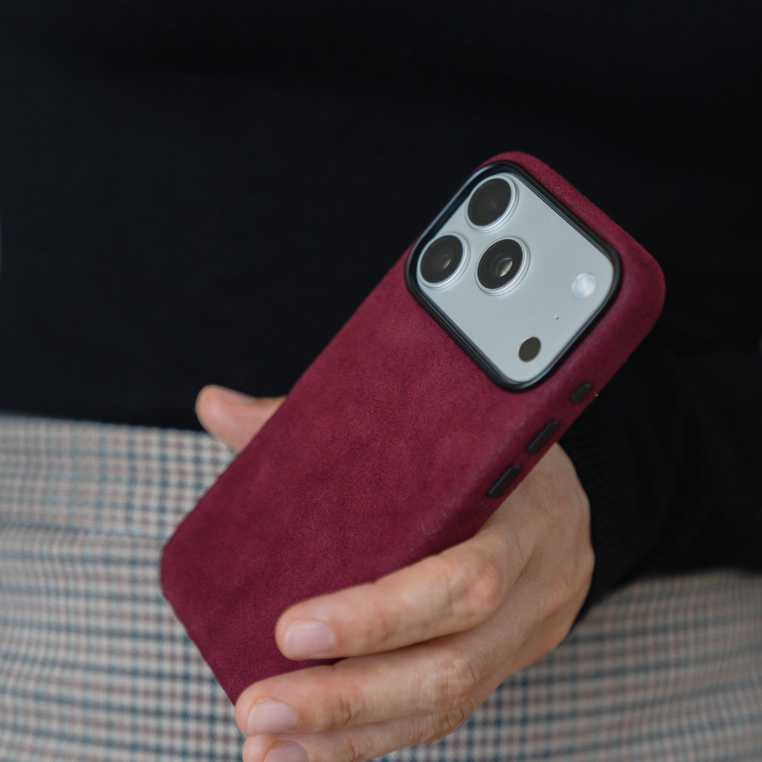 In der Hand ein arktis iPhone 17 Pro Hülle aus Alcantara in kastanienbraun, bekleidet mit einem schwarzen Oberteil und einem karierten Rock.