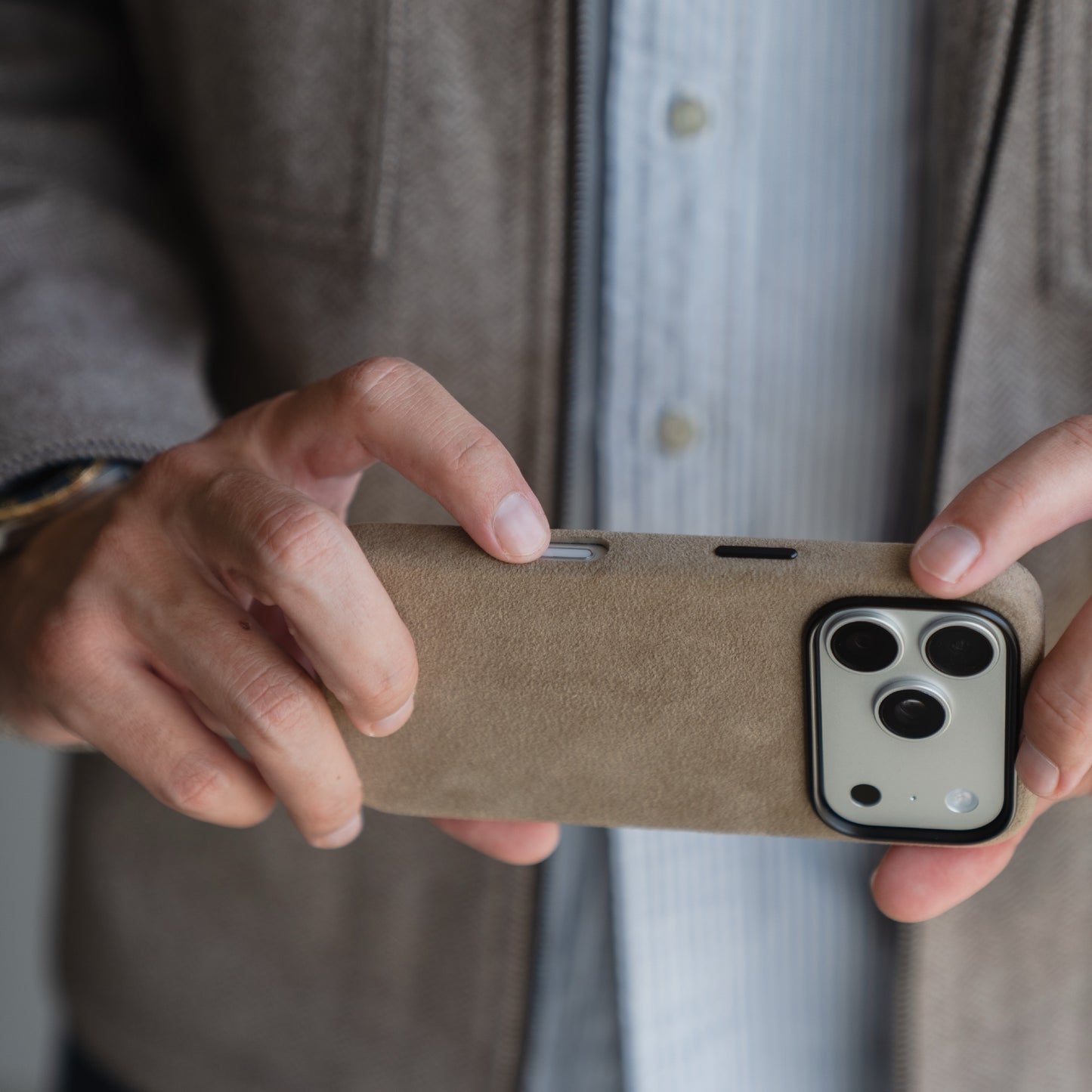Eine Person hält ein Smartphone mit einer arktis iPhone 17 Pro Hülle aus Alcantara in Beige, trägt eine graue Jacke und ein gestreiftes Hemd. Diese elegante Hülle von arktis wird mit Sorgfalt gefertigt und ist stolz Made in Italy.