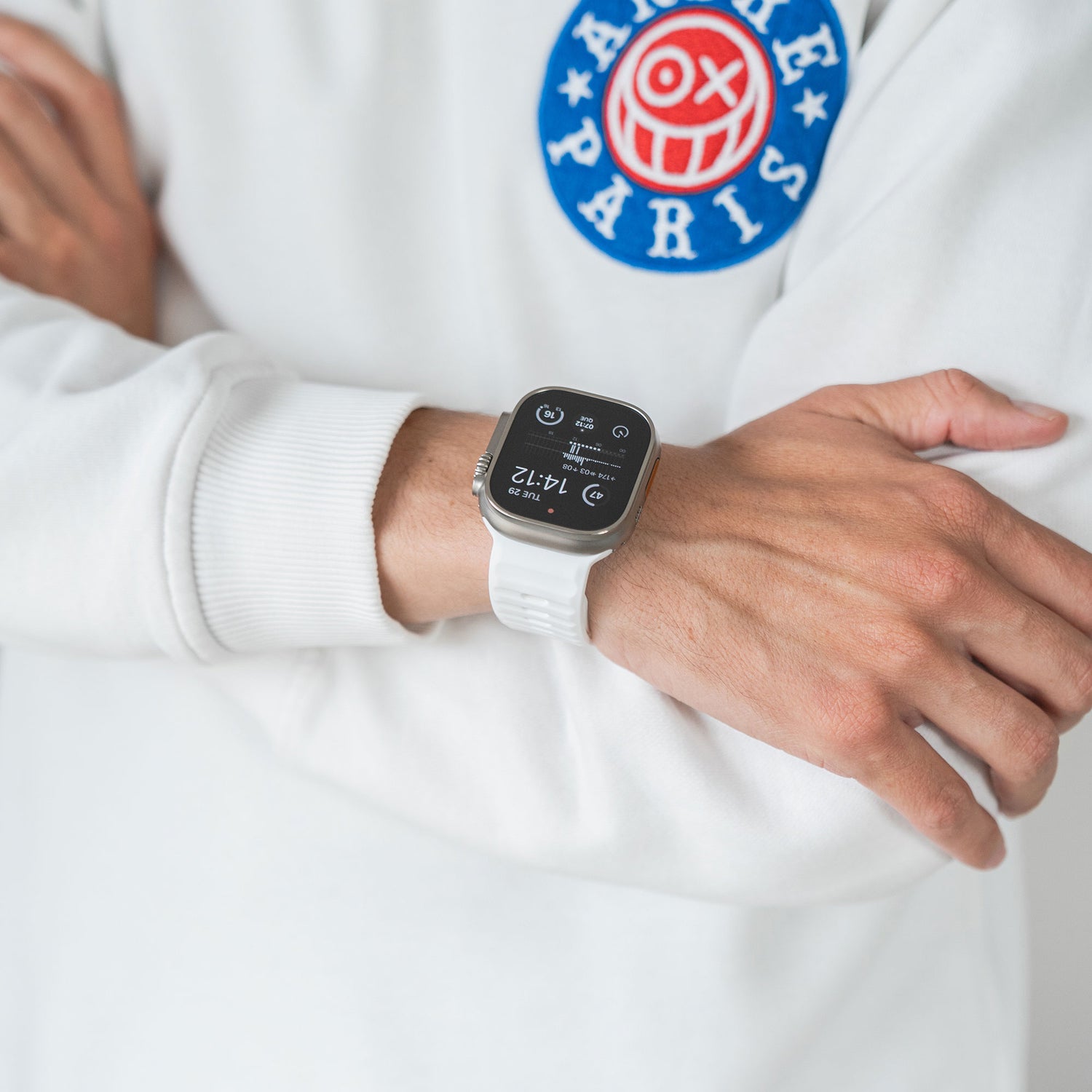 Eine Person verschränkt die Arme und trägt ein weißes Sweatshirt mit einem blau-rot-weißen Brustaufnäher. Am Handgelenk trägt sie das arktisband Apple Watch Silikonarmband „Active“ von arktisband, das die Uhrzeit und Fitnessdaten anzeigt.