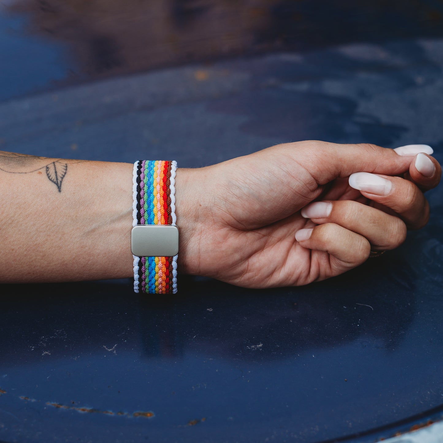 Eine Hand mit langen, manikürten Nägeln und einem kleinen Tattoo trägt das arktisband Geflochtene Apple Watch Armband „Casual“ Rainbow Edition von arktisband mit Regenbogenstreifen und silbernem Verschluss, der auf einer dunkelblauen Oberfläche ruht.