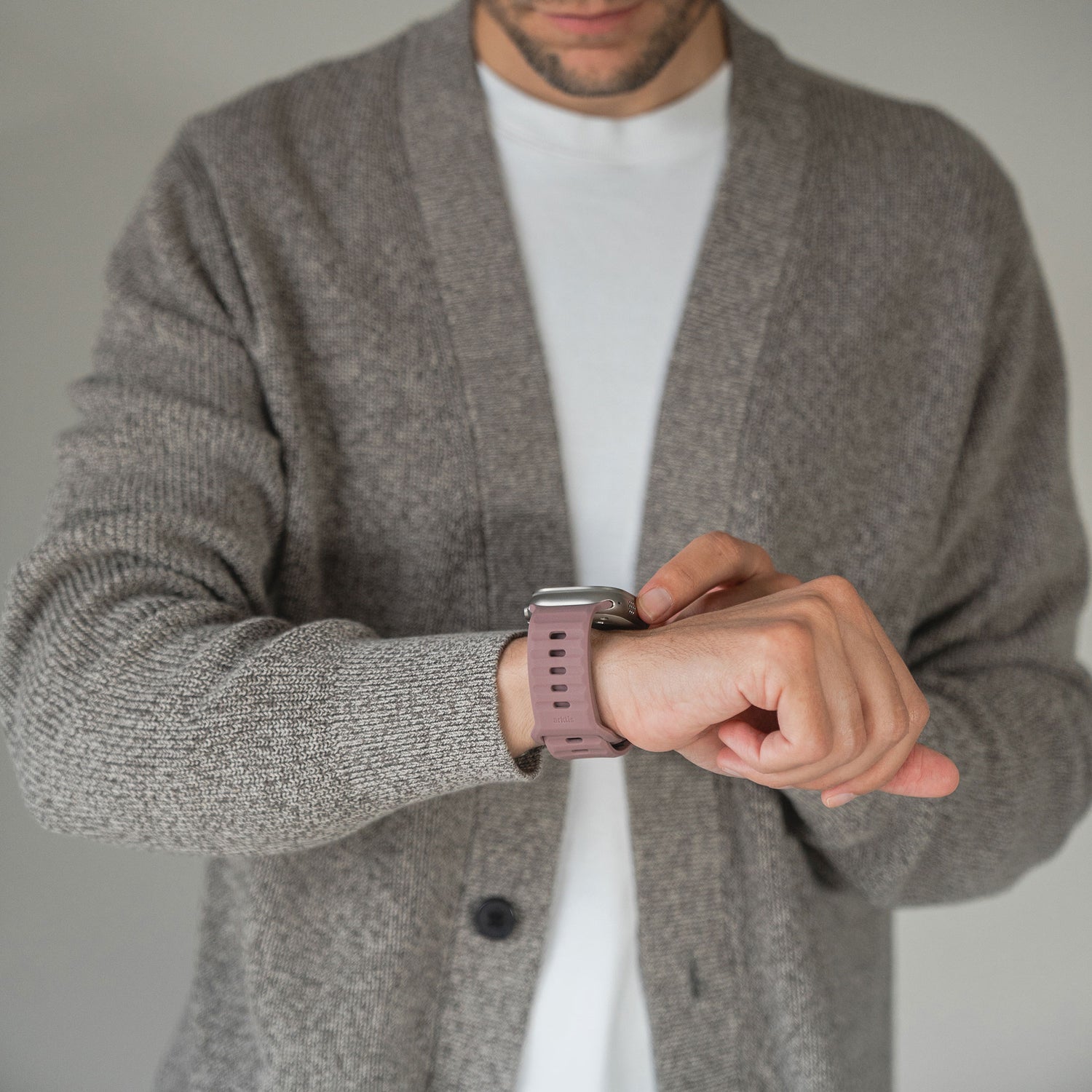 Eine Person in einer grauen Strickjacke und einem weißen Hemd blickt auf ein Arktisband Apple Watch Silikonarmband „Active“ an ihrem linken Handgelenk und berührt es, wobei ihr Gesicht teilweise außerhalb des Bildes ist.