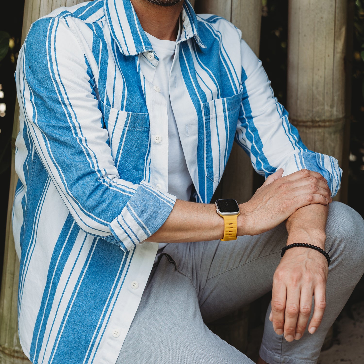 Ein Mann in einem blau-weiß gestreiften Hemd, einem weißen T-Shirt und einer hellgrauen Hose sitzt mit einem Arm auf seinem Knie und trägt ein arktisband Apple Watch Silikonarmband „Active“ von arktisband in Gelb und ein schwarzes Perlenarmband.