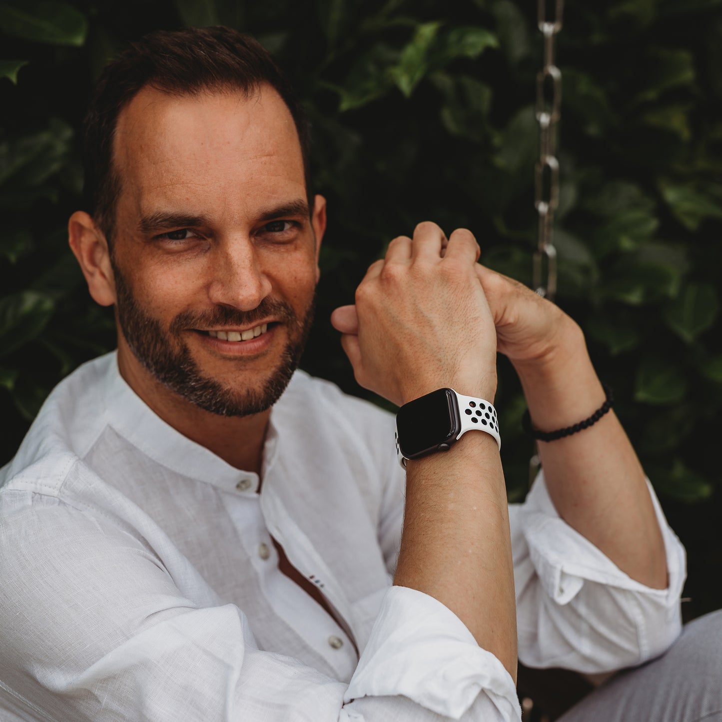 Ein Mann im weißen Hemd sitzt im Freien vor grünem Laub und lächelt in die Kamera. Er hat kurze dunkle Haare, einen Bart und trägt ein Arktisband Apple Watch Silikonarmband „Energy“ am linken Handgelenk.