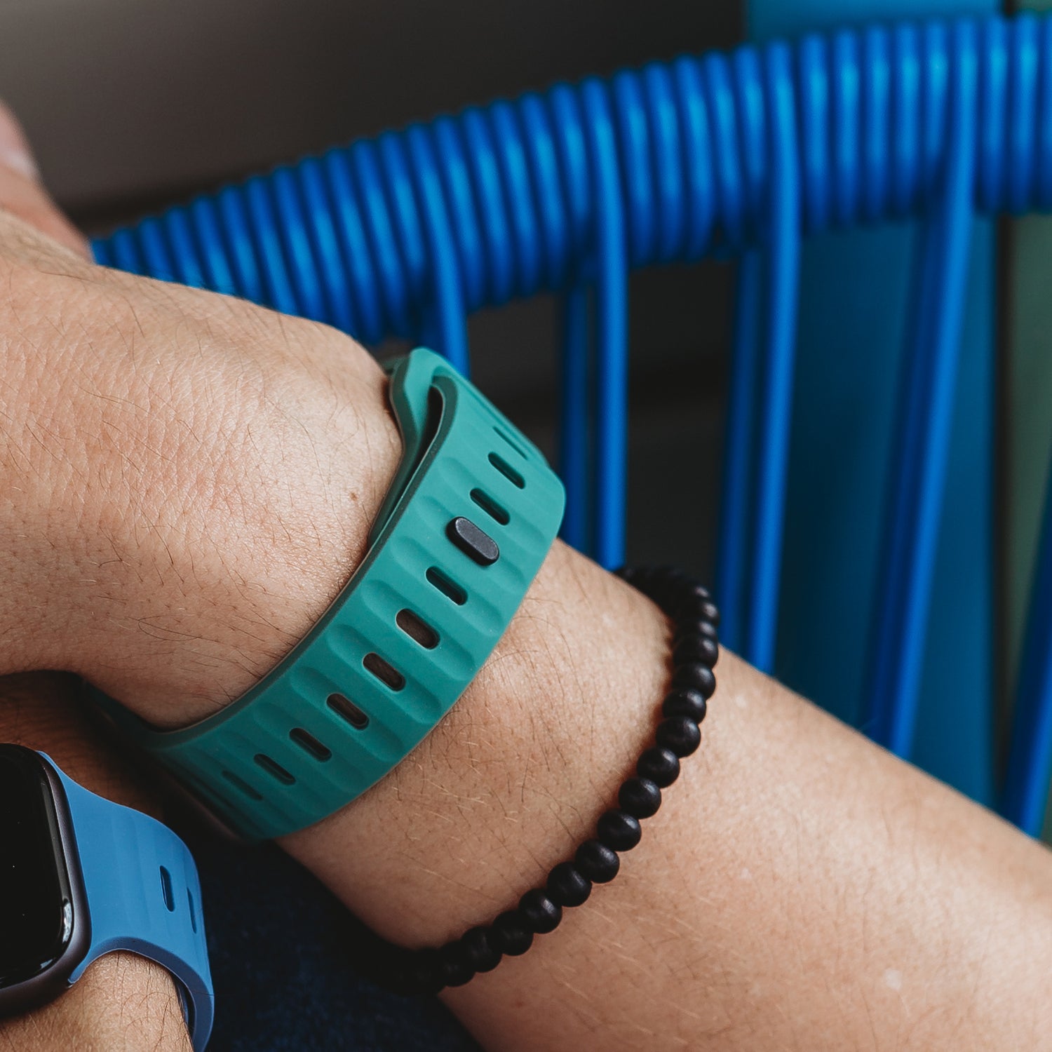 Eine Nahaufnahme eines Handgelenks mit einem grünen Arktisband Apple Watch Silikonarmband „Active“, einem blauen Apple Watch-Armband und einem schwarzen Perlenarmband, mit einem blauen Stuhl im Hintergrund.