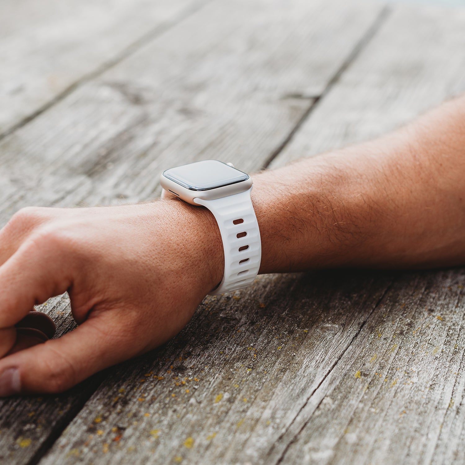 Das linke Handgelenk einer Person trägt das arktisband Apple Watch Silikonarmband „Active“ in Weiß mit atmungsaktivem Design von arktisband und ruht auf einer verwitterten Holzoberfläche im Freien. Der Bildschirm der Smartwatch bleibt ausgeschaltet.
