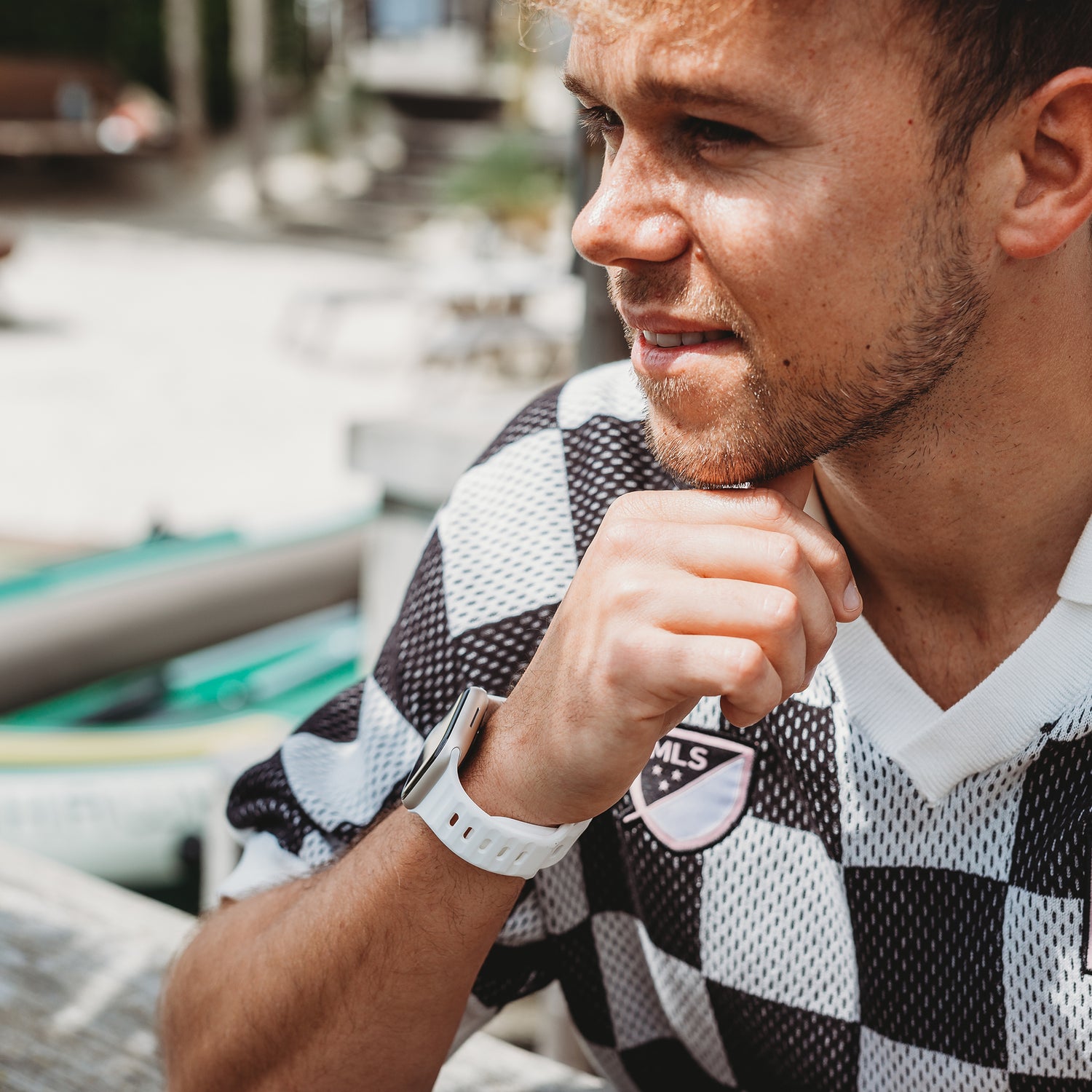 Ein Mann in einem schwarz-weiß karierten Hemd mit MLS-Abzeichen sitzt im Freien und stützt sein Kinn auf seine Hand. Er trägt das arktisband Apple Watch Silikonarmband „Active“ von arktisband. Im Hintergrund sind verschwommene Kajaks zu sehen.