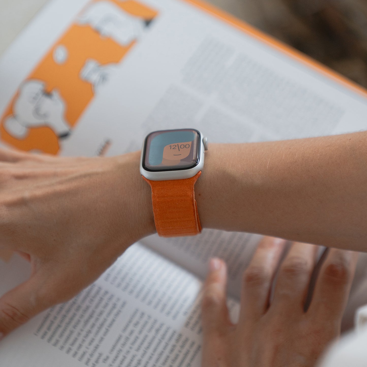 Jemand, der ein orangefarbenes Arktisband Apple Watch magnetisches Armband aus Alcantara trägt, liest ein aufgeschlagenes Buch mit orangefarbenen und schwarzen Illustrationen.