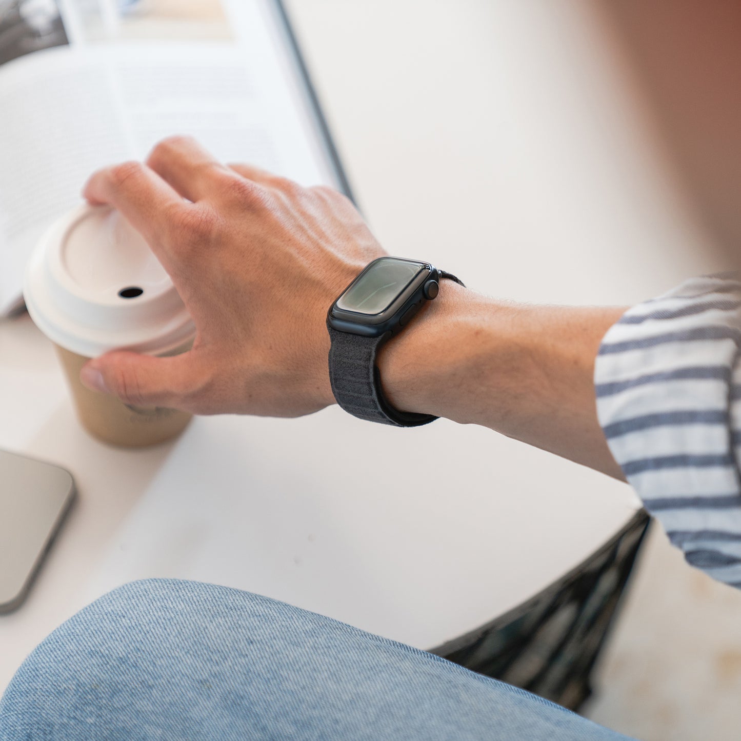 Eine Person, die die arktisband Apple Watch magnetisches Armband aus Alcantara von arktisband trägt, hält eine Kaffeetasse neben einer aufgeschlagenen Zeitschrift und fängt echtes italienisches Lebensgefühl ein.