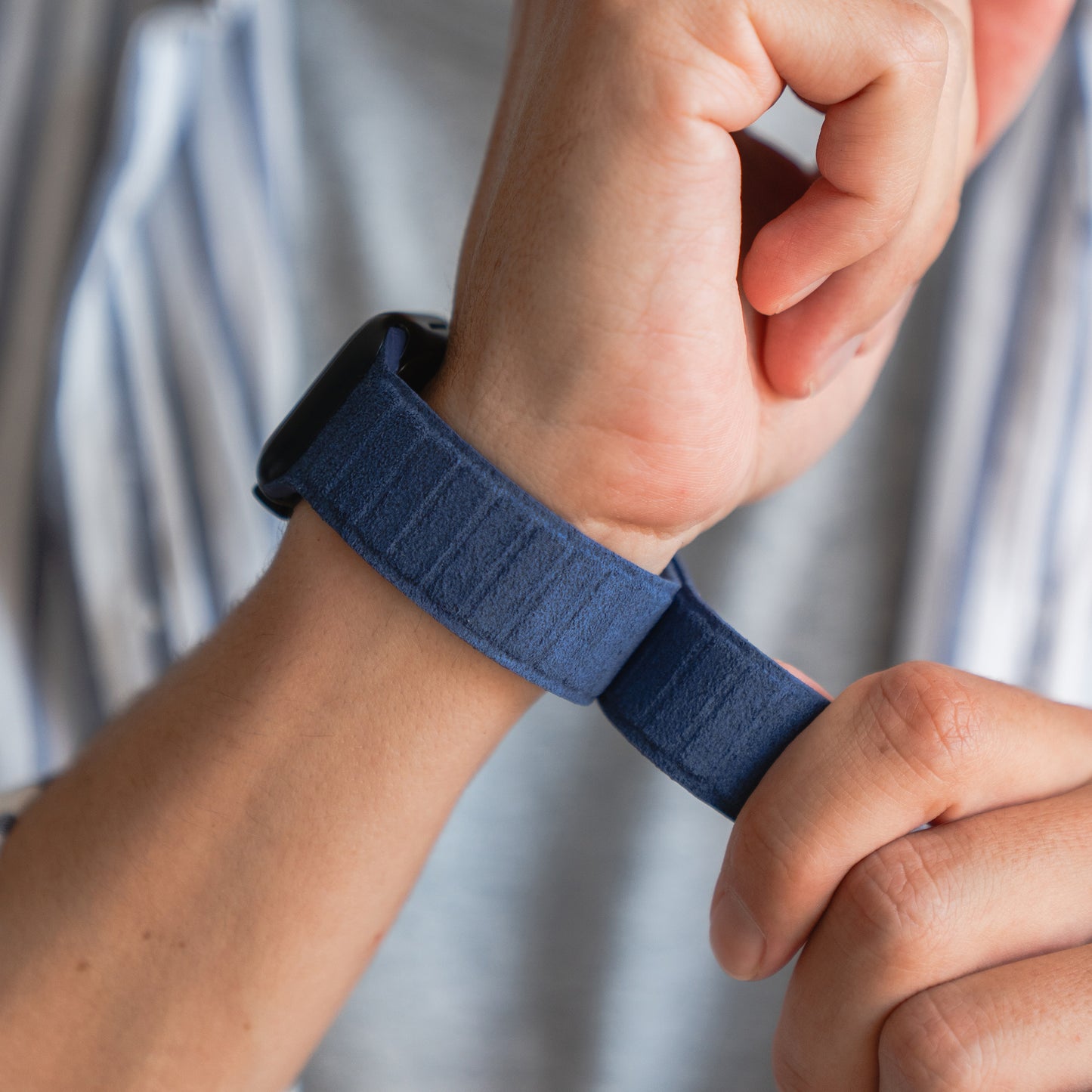 Jemand befestigt die blaue arktisband Apple Watch magnetisches Armband aus Alcantara von arktisband an seinem Handgelenk und trägt dabei ein hellgrau gestreiftes Hemd, das echtes italienisches Lebensgefühl einfängt.