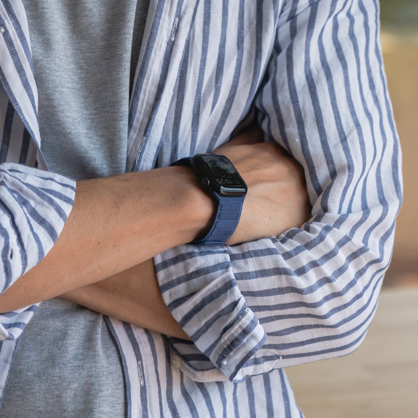 Eine Person in einem gestreiften Hemd trägt das Arktisband Apple Watch magnetisches Armband aus Alcantara und strahlt mit verschränkten Armen italienisches Lebensgefühl aus.