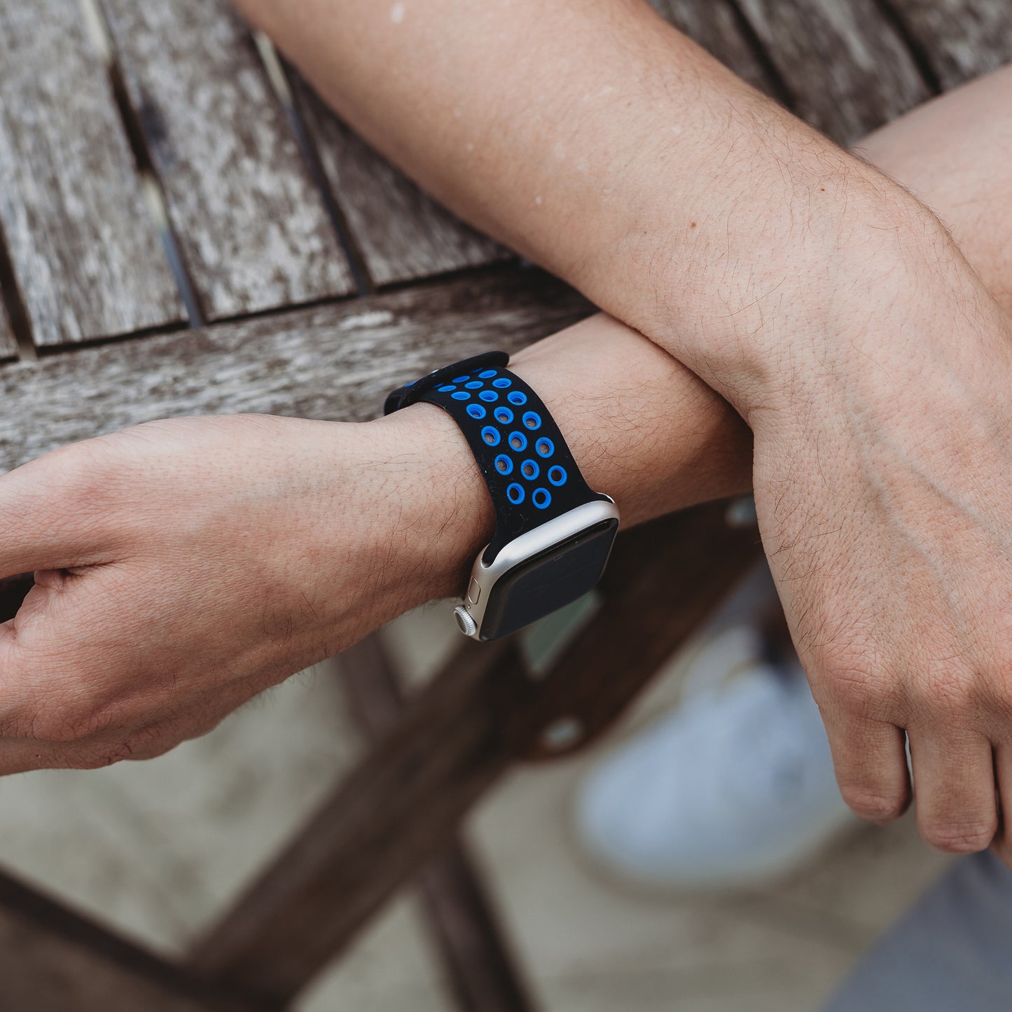 Jemand, der ein arktisband Apple Watch Silikonarmband „Energy“ in Schwarz und Blau von arktisband trägt, legt seine Arme auf einen verwitterten Holztisch, im Hintergrund ist ein Teil seines weißen Schuhs zu sehen.