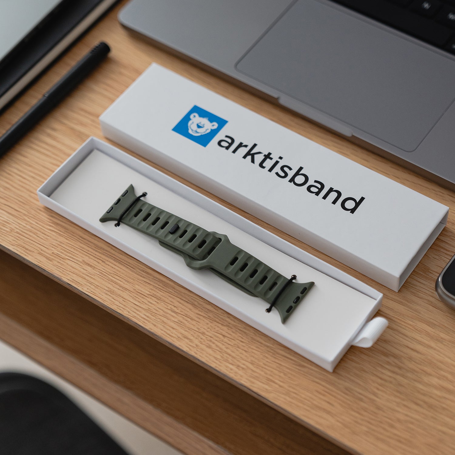 Das arktisband Apple Watch Silikonarmband „Active“ in Grün, bekannt für sein atmungsaktives Design, wird in einer offenen weißen arktisband-Box auf einem Holzschreibtisch neben einem Laptop, einem Stift und einem Tablet gezeigt.
