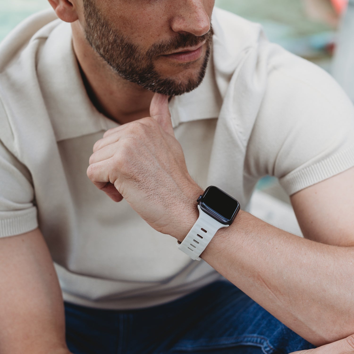 Ein Mann in einem kurzärmeligen beigen Poloshirt und Bluejeans stützt sein Kinn auf seine Hand und trägt ein arktisband Apple Watch Silikonarmband „Active“ von arktisband; das weiße, atmungsaktive Band umschließt eine Smartwatch mit ausgeschaltetem Bildschirm.