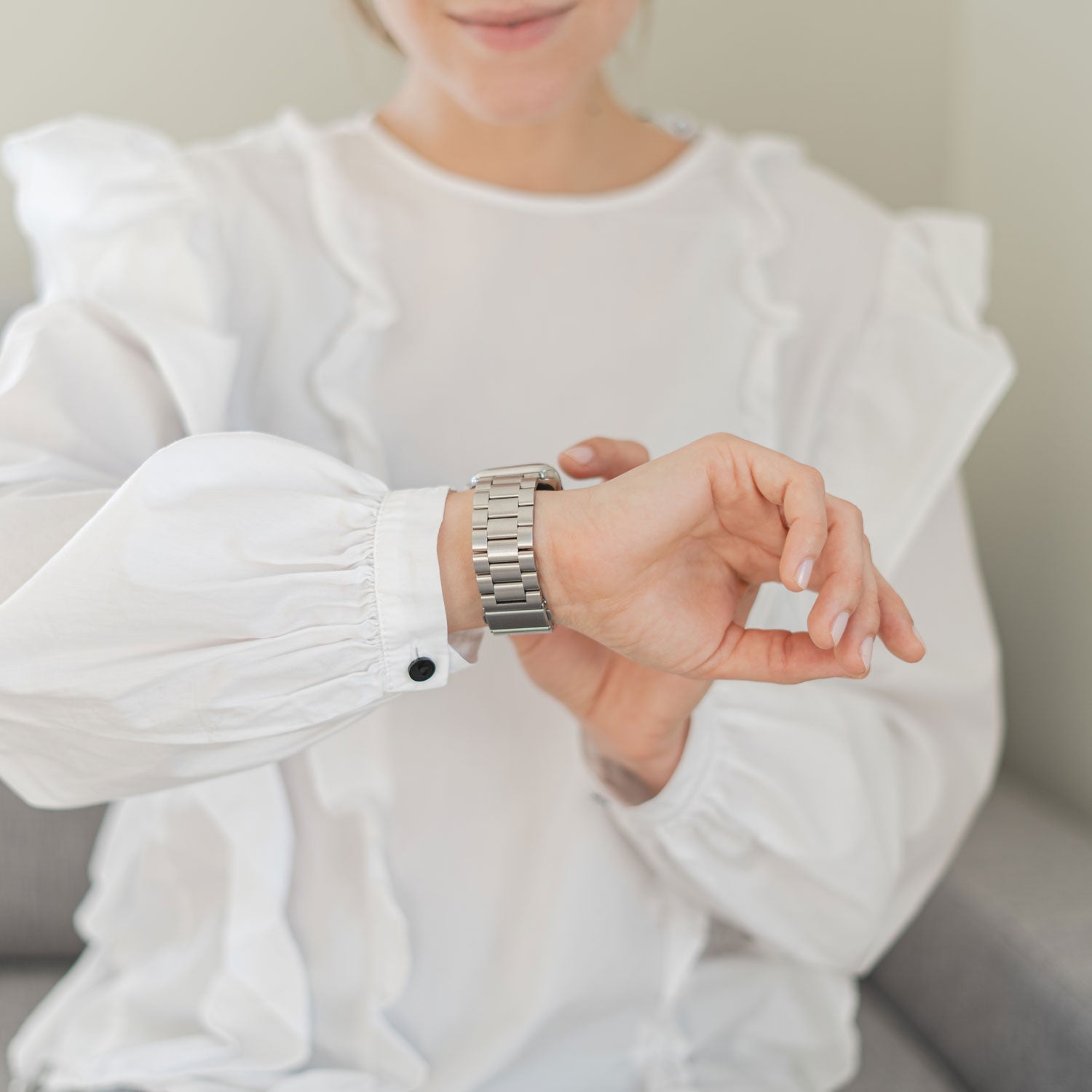 Eine Person in einer weißen Rüschenbluse rückt ihr arktisband Apple Watch Gliederarmband „Arctica“ von arktisband zurecht und blickt dabei auf ihre Armbanduhr; nur die untere Hälfte ihres Gesichts ist zu sehen.